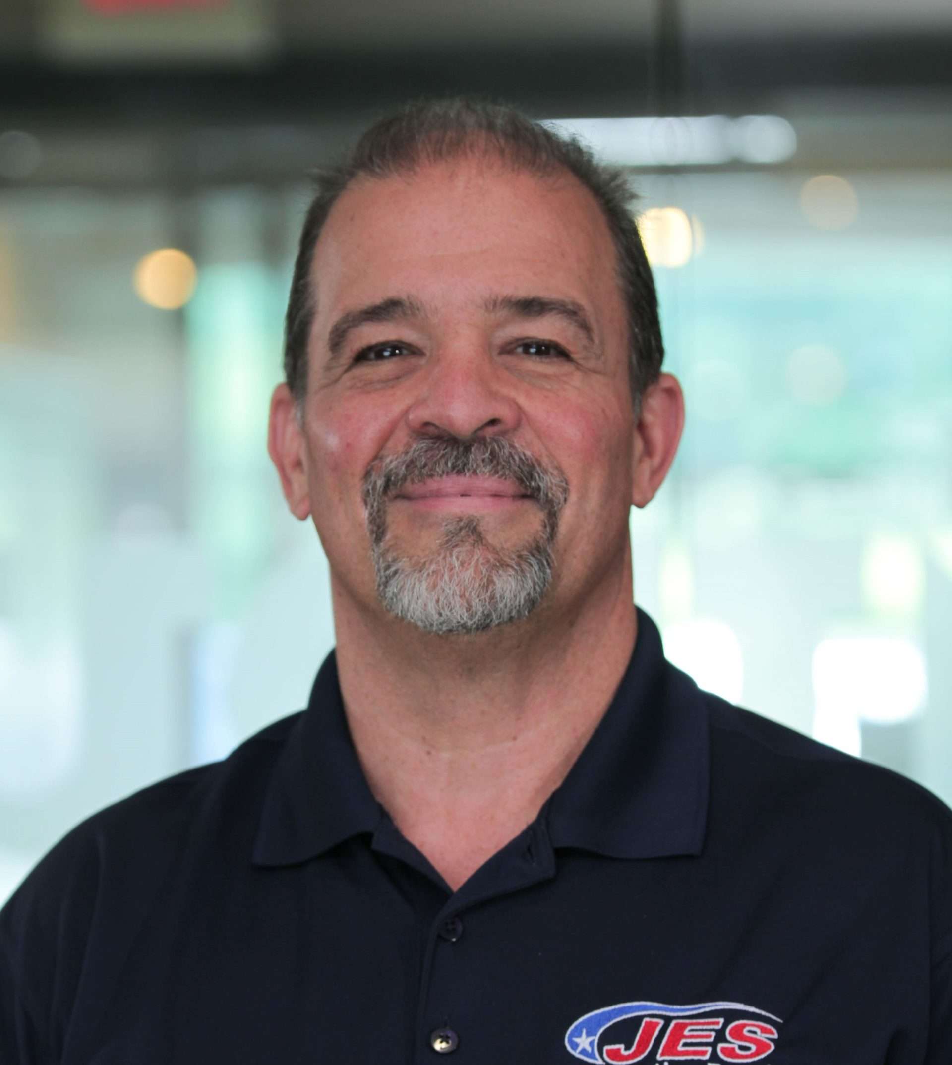 A man with a goatee smiles, wearing a dark polo shirt with a "JES" logo. Blurred background with soft lighting.
