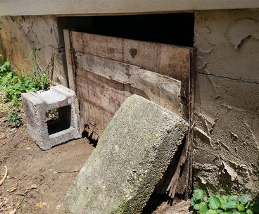 A concrete block and wooden board partially cover a hole in a building’s foundation.