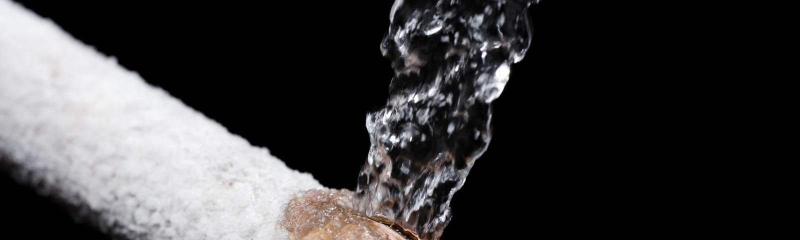 Water bursts from a frozen pipe covered in frost against a black background.