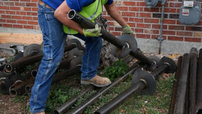 Worker in jeans and safety vest lifting metal pipes outdoors near a brick wall, prepping for foundation repair.