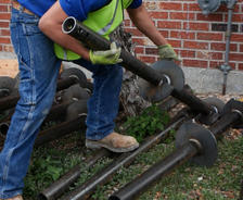 Worker in jeans and safety vest lifting metal pipes outdoors near a brick wall, prepping for foundation repair.