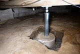 A sturdy support beam rests in the crawl space, held firm by a steel jack and securely anchored on a robust concrete footing.