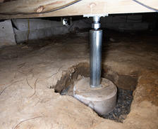 A sturdy support beam rests in the crawl space, held firm by a steel jack and securely anchored on a robust concrete footing.