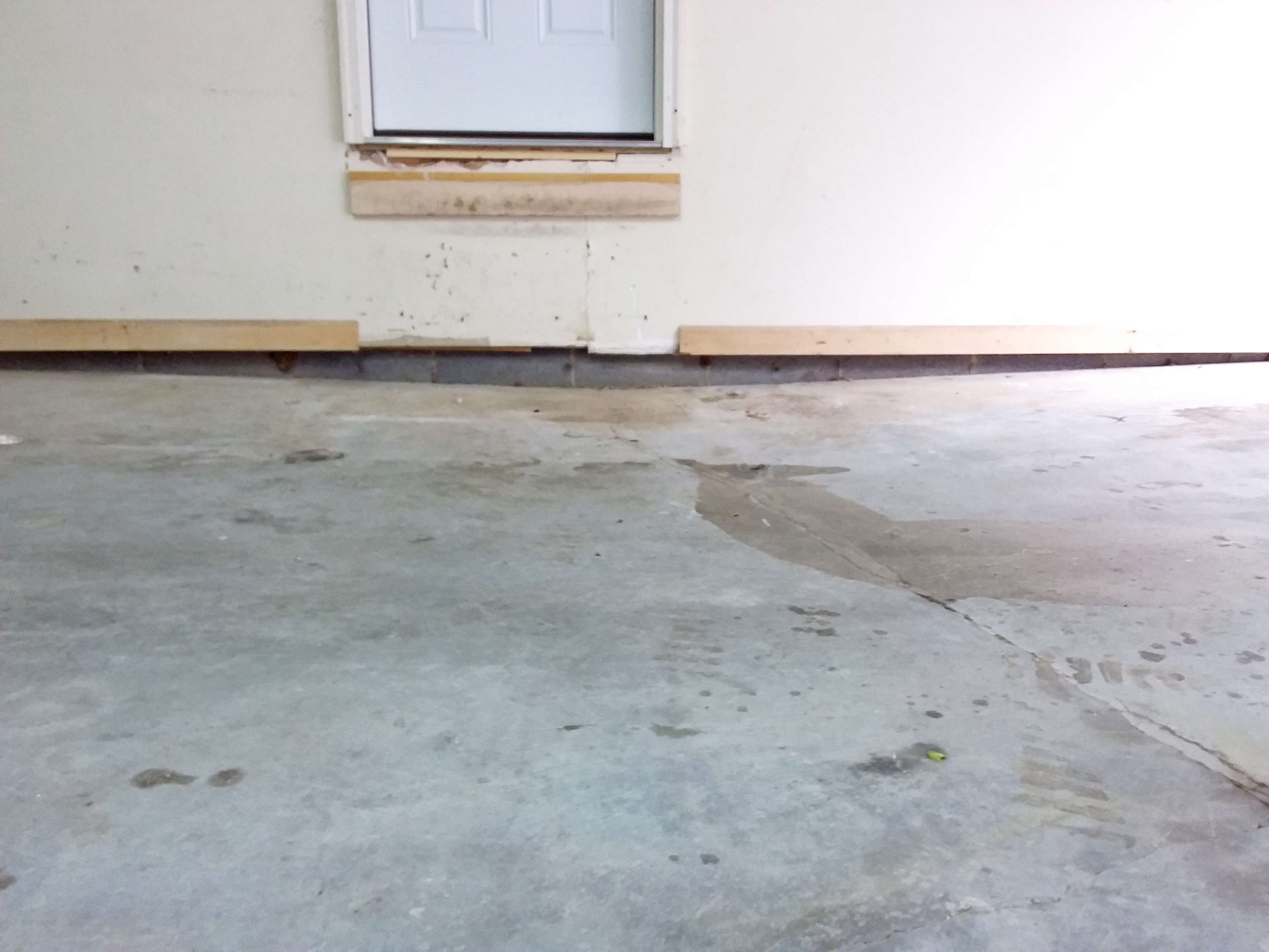 Concrete garage floor with water stains near a wall and door, wood boards attached to the wall.