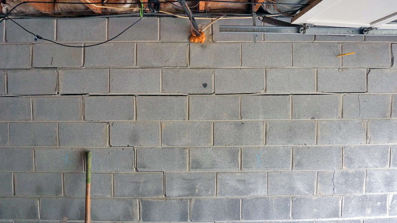 Cinder block wall with visible horizontal and stair-step cracks, exposed wires, and a broom handle.
