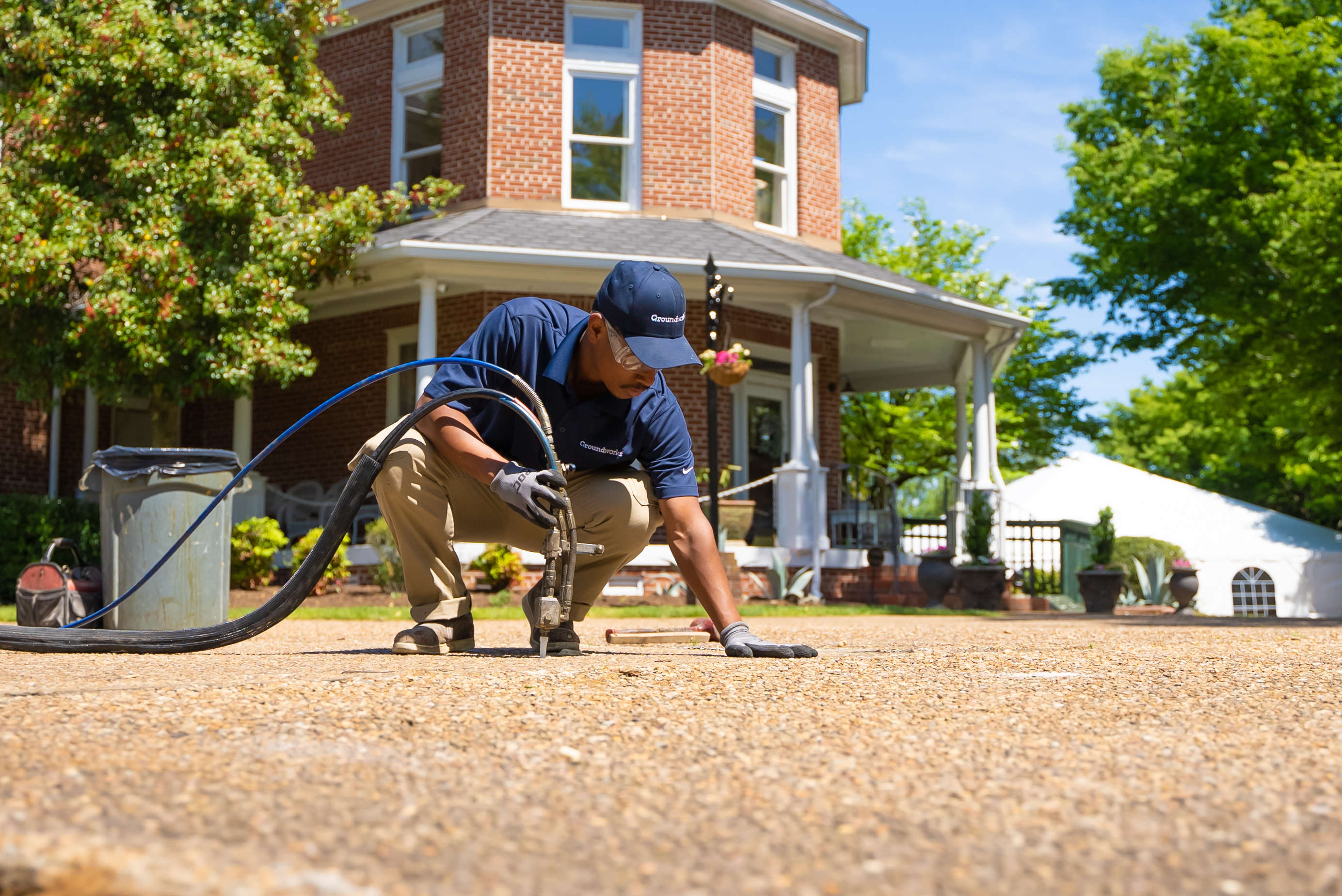 Worker in blue shirt kneels on driveway using equipment, large brick house and trees in background.