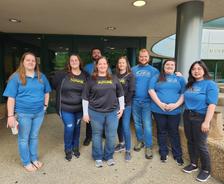 A group of nine people stand and smile together outside a building, wearing matching blue and black shirts.