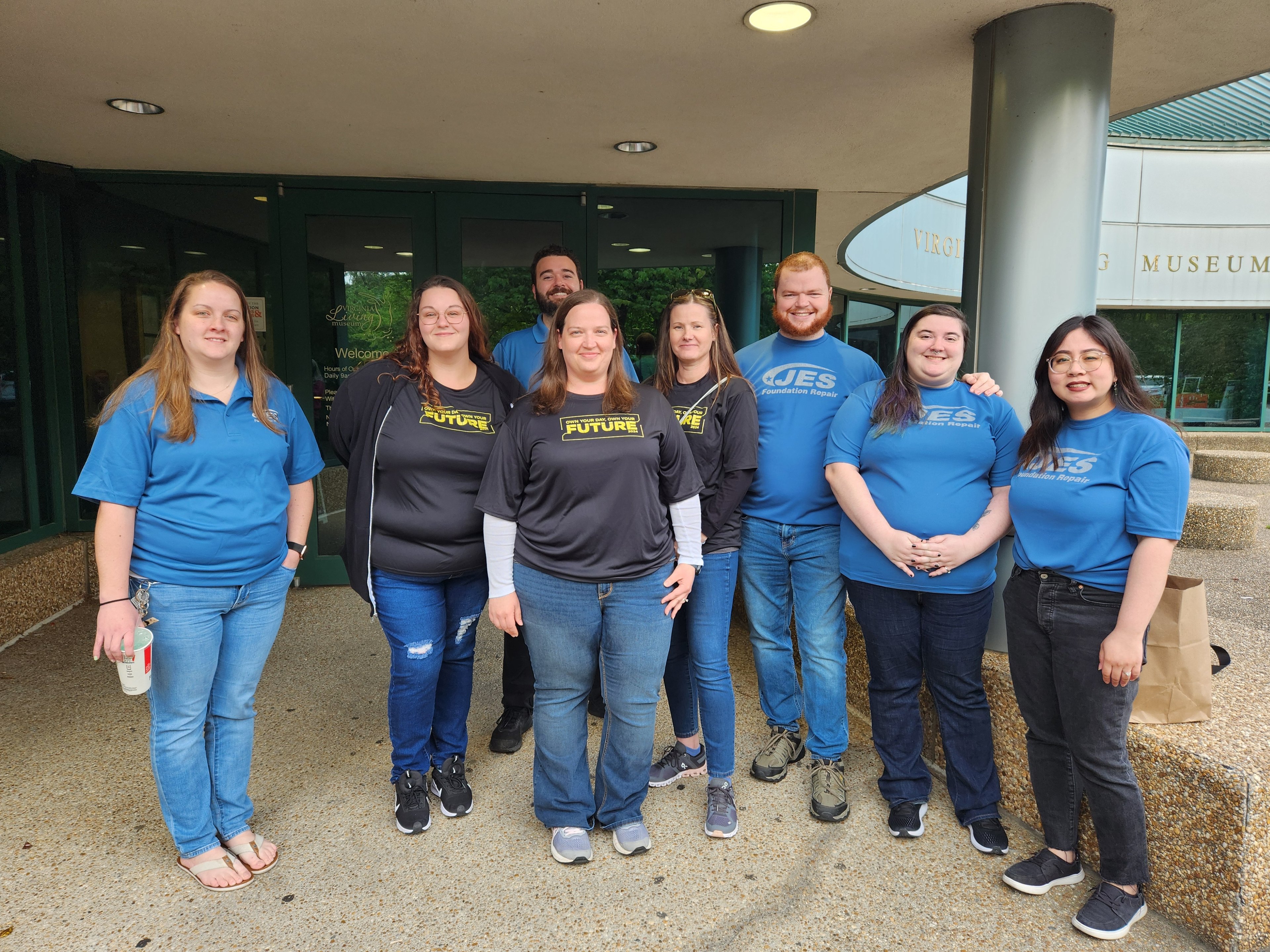 A group of nine people stand and smile together outside a building, wearing matching blue and black shirts.