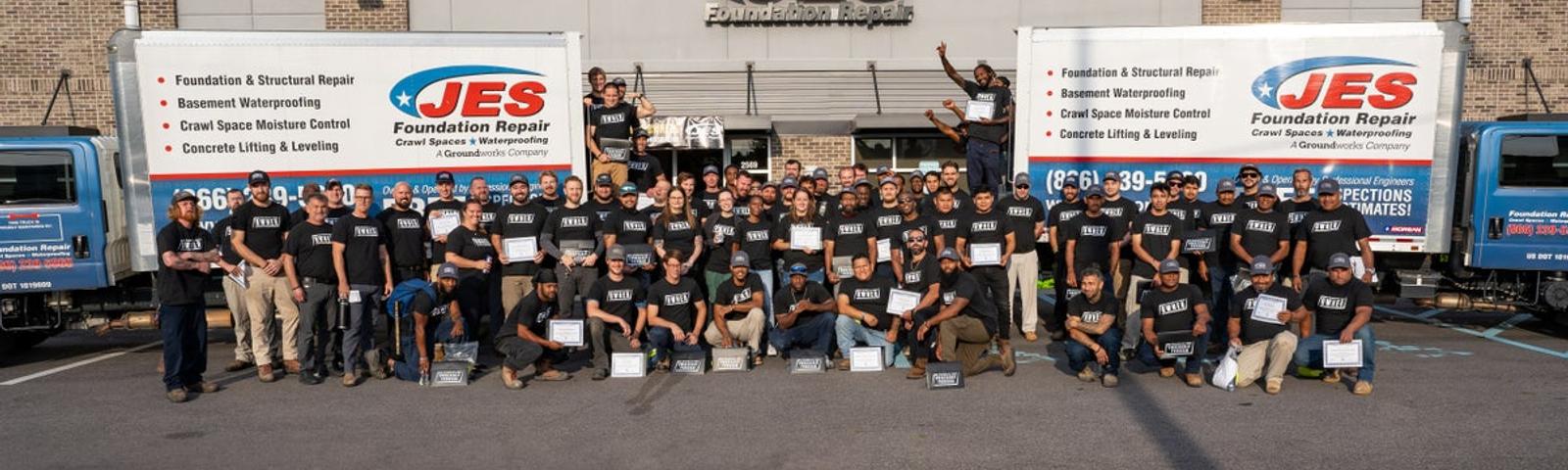 A large group of people in black shirts pose in front of trucks outside a building with a JES Foundation Repair sign.