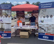 Outdoor booth with banners about basement and foundation solutions, staffed by a person under a red canopy.