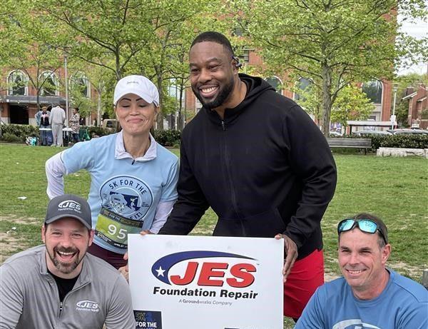 Four people posing with a JES Foundation Repair sign at a park event.