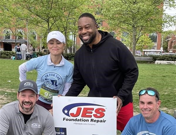 Four people posing with a JES Foundation Repair sign at a park event.