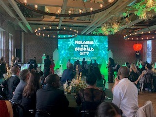 A formal sponsorship event with JES Foundation Repair team members seated at tables, featuring a large screen that reads "Welcome to the Emerald City Kaleidoscope Ball.
