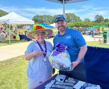 Two people smiling, one wearing a colorful hat, holding a gift basket at an outdoor fair or market.