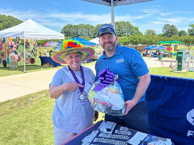Two people smiling, one wearing a colorful hat, holding a gift basket at an outdoor fair or market.