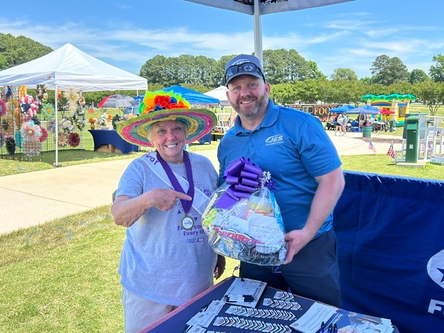 Two people smiling, one wearing a colorful hat, holding a gift basket at an outdoor fair or market.
