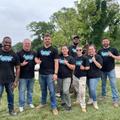 Eight JES Foundation Repair team members in matching shirts stand together outdoors, smiling at the camera for Habitat for Humanity volunteer efforts.