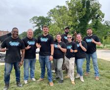 Eight JES Foundation Repair team members in matching shirts stand together outdoors, smiling at the camera for Habitat for Humanity volunteer efforts.