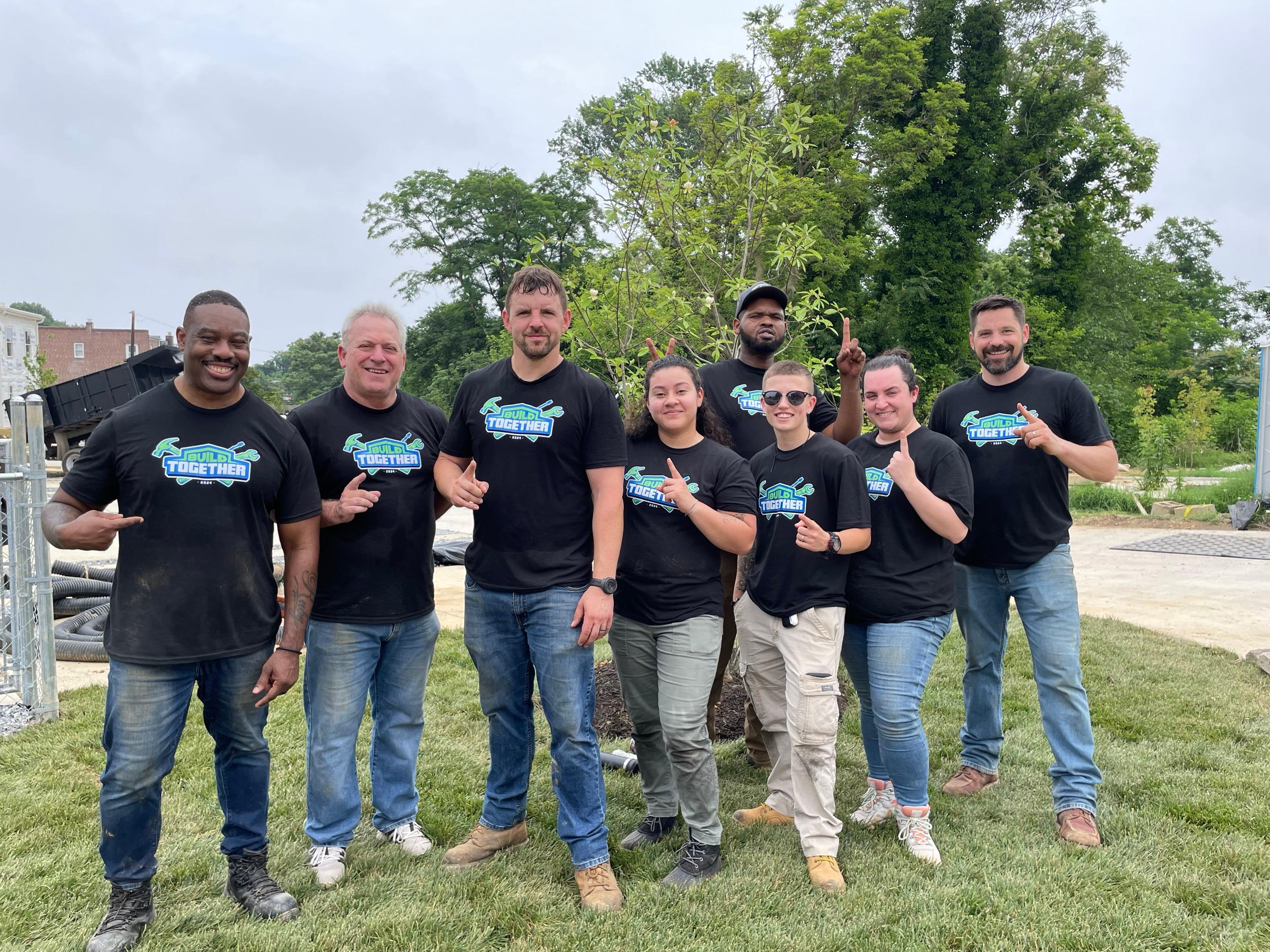 Eight JES Foundation Repair team members in matching shirts stand together outdoors, smiling at the camera for Habitat for Humanity volunteer efforts.