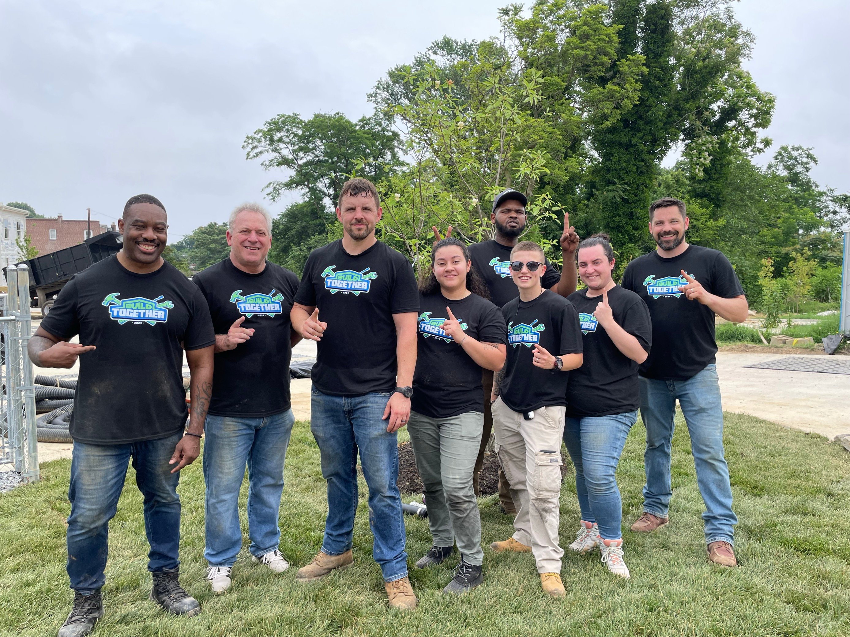 Eight JES Foundation Repair team members in matching shirts stand together outdoors, smiling at the camera for Habitat for Humanity volunteer efforts.