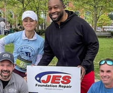Four people posing with a "JES Foundation Repair" sign at an outdoor event, with trees and people in the background.