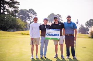 Four men on a golf course holding a "2024 Golf Outing" sign, smiling on a sunny day.