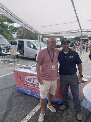Two JES Foundation Repair team members smiling under a canopy at McShin Foundation Recovery event near a table covered with a company-branded cloth.