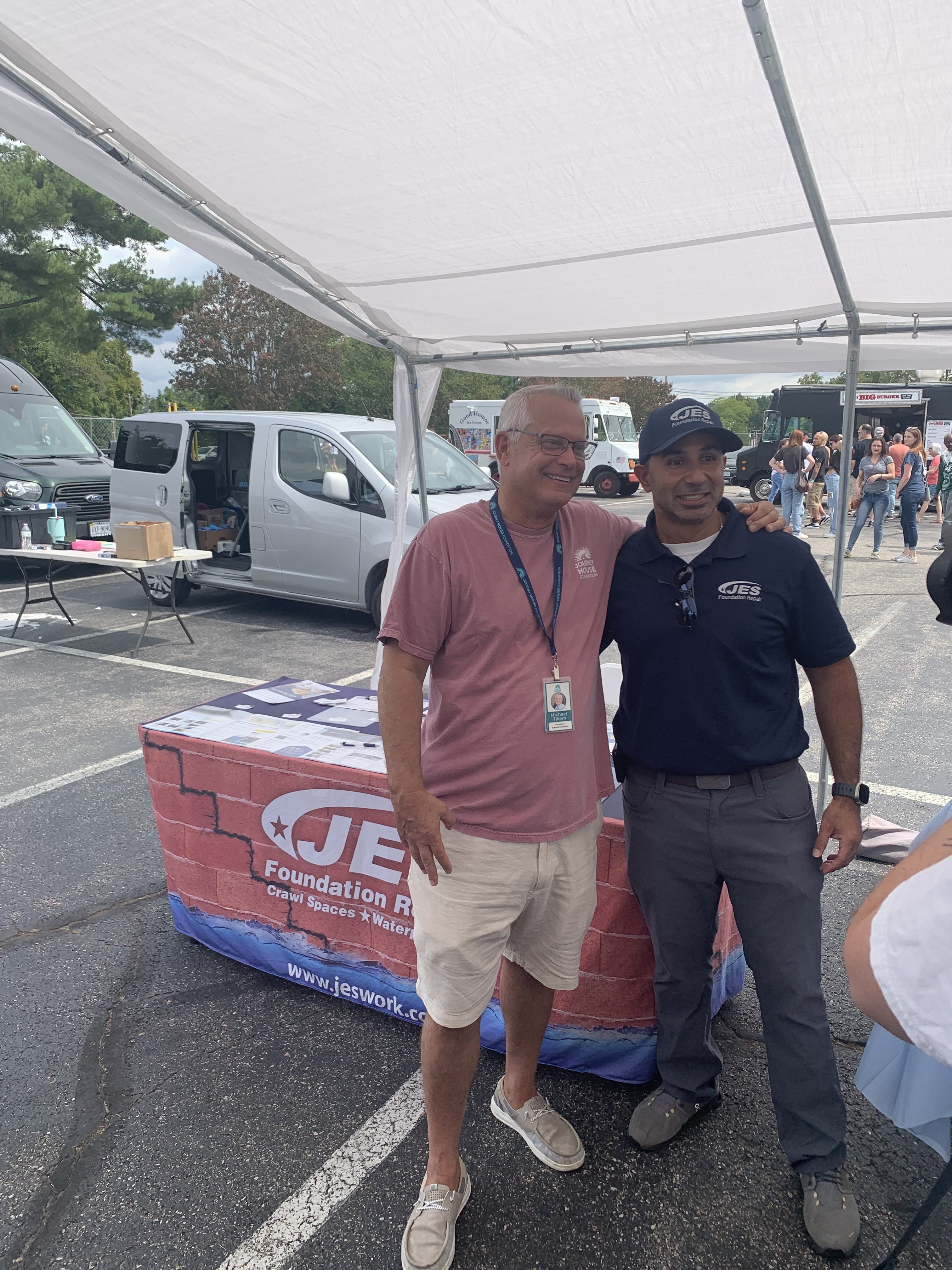 Two JES Foundation Repair team members smiling under a canopy at McShin Foundation Recovery event near a table covered with a company-branded cloth.