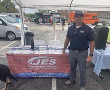 A man in a JES shirt stands by a JES Foundation Repair booth at an outdoor event.