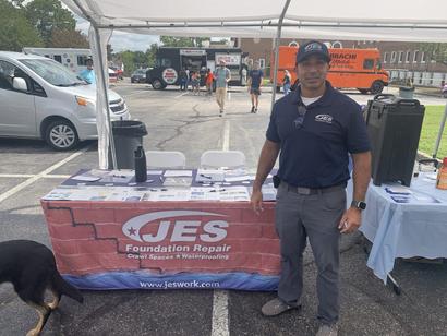 A man in a JES shirt stands by a JES Foundation Repair booth at an outdoor event.