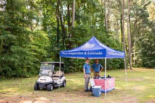Two people at an outdoor booth with a tent offering free inspections, near a golf cart, in a wooded area.
