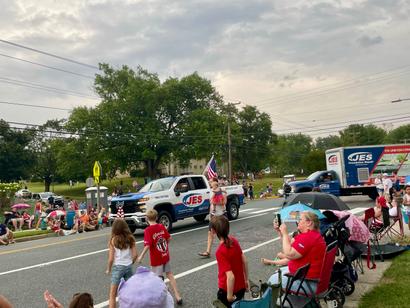 Parade scene with a truck, people waving, and spectators lined along a street with trees in the background.