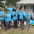 Eight JES Foundation Repair volunteers in blue Habitat for Humanity shirts pose and flex arms in front of a house repair site.