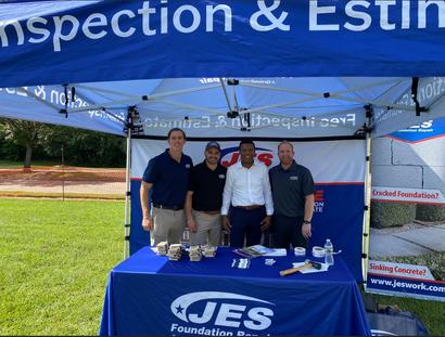 Four men stand smiling under a JES Foundation Repair tent at an outdoor event with promotional materials on the table.