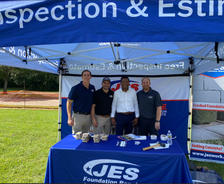 Four men stand smiling under a JES Foundation Repair tent at an outdoor event booth on a sunny day at National Disaster Preparedness event.