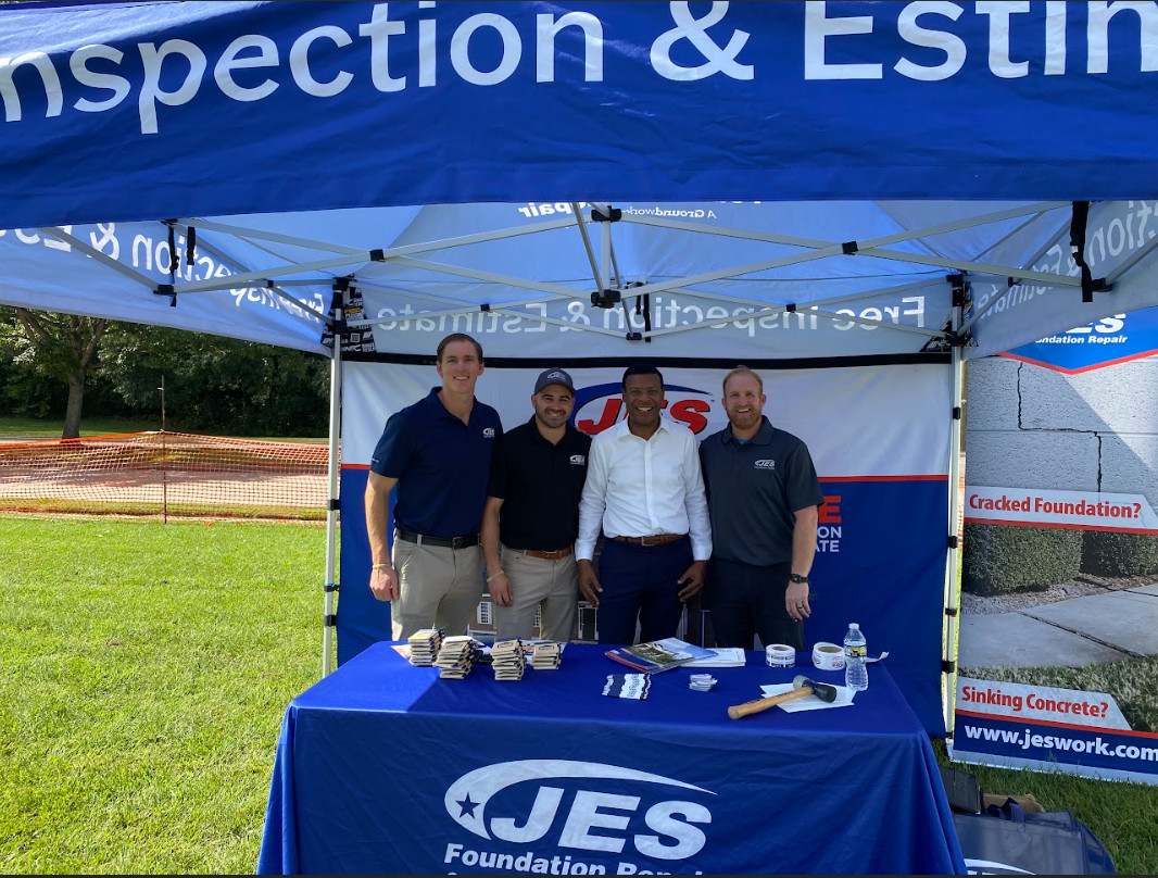 Four men stand smiling under a JES Foundation Repair tent at an outdoor event booth on a sunny day at National Disaster Preparedness event.