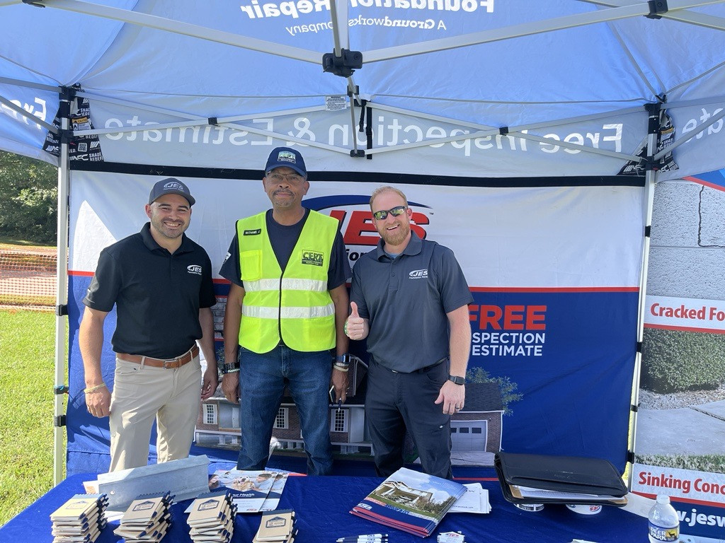 Three men smile under a promotional tent, one gives a thumbs up, with brochures displayed on the table.