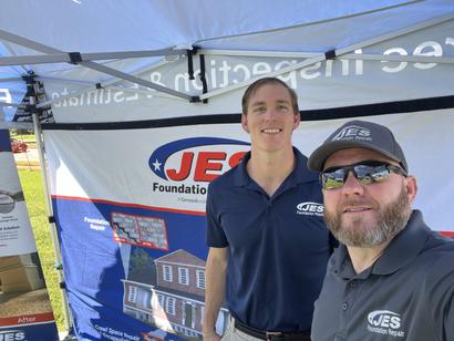 Two men in JES Foundation shirts stand smiling at a company booth under a branded tent at an outdoor event.