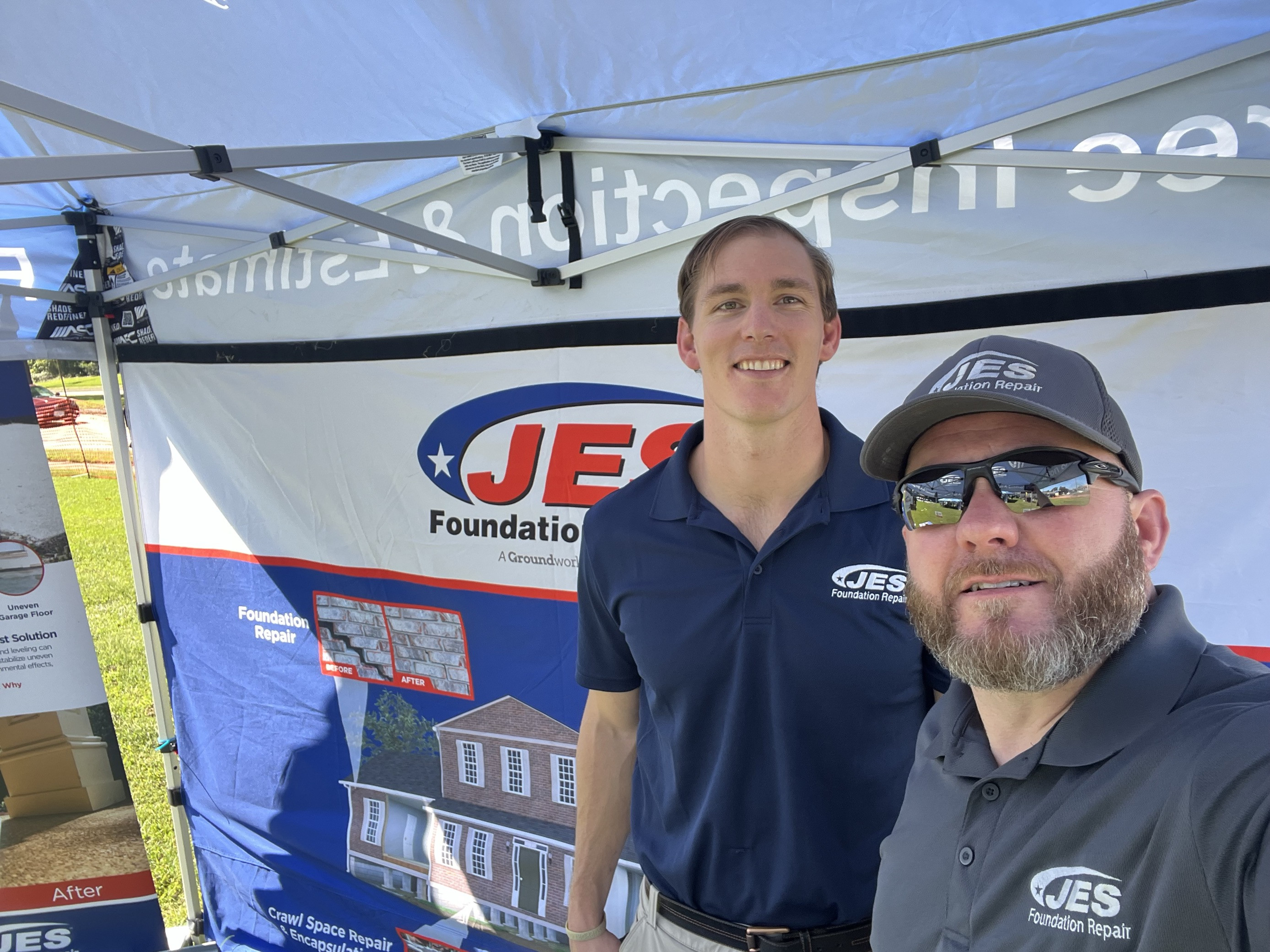 Two men in JES Foundation Repair shirts stand under a promotional tent at an outdoor event.