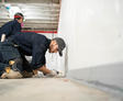 Two workers kneel on the floor, applying sealant along a white wall in a construction area for basement sealing and waterproofing.