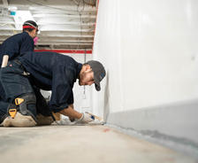 Two workers kneel on the floor, applying sealant along a white wall in a construction area for basement sealing and waterproofing.