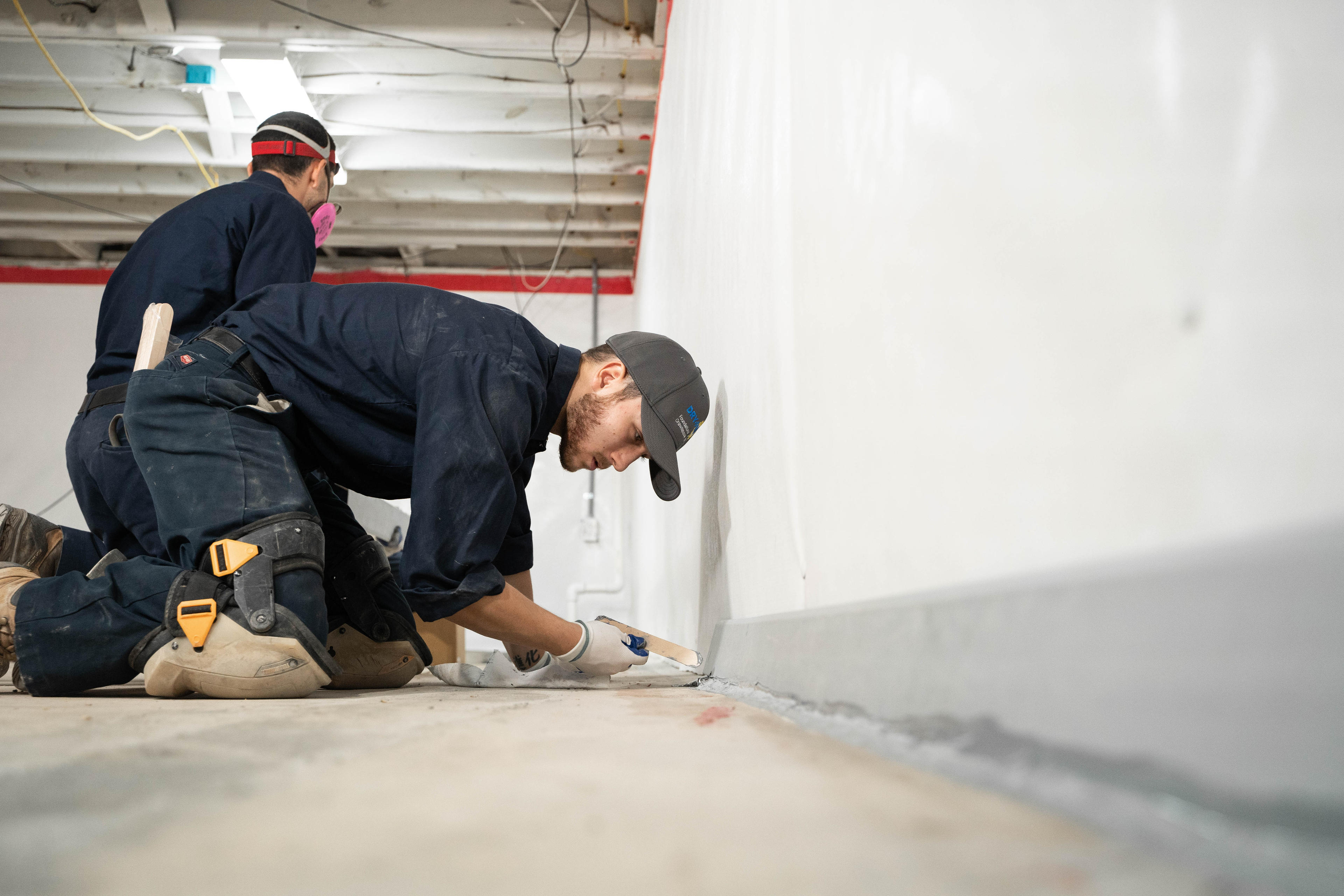Two workers kneel on the floor, applying sealant along a white wall in a construction area for basement sealing and waterproofing.