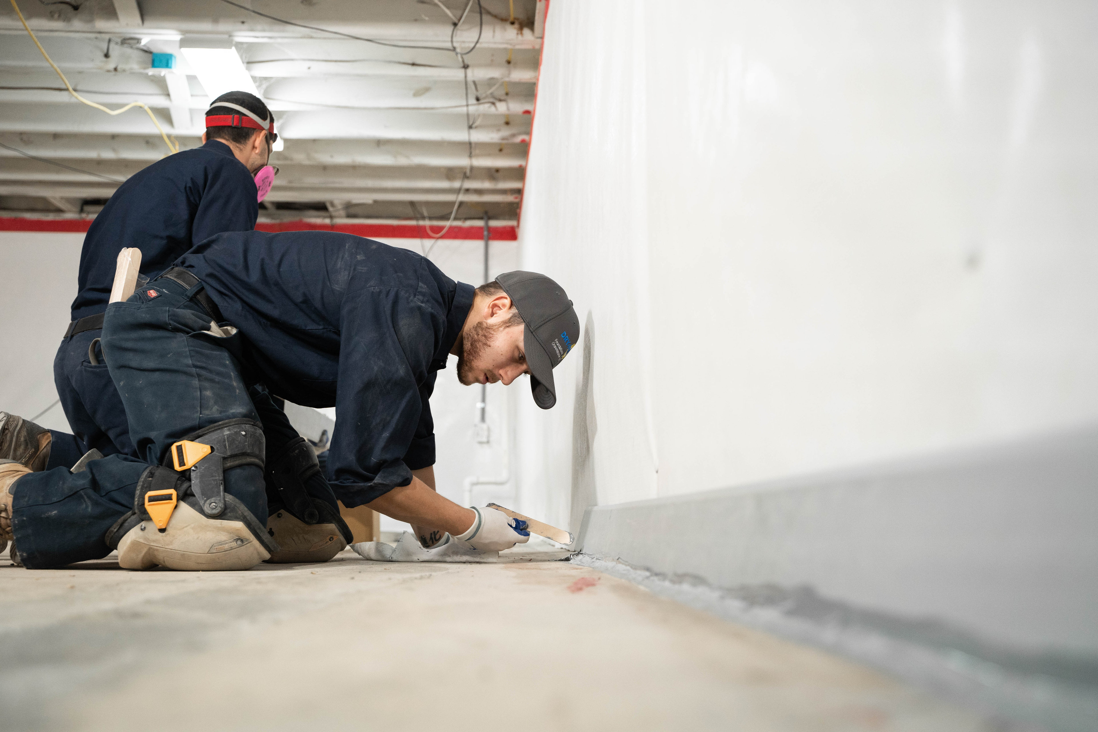 Two workers kneel on the floor, applying sealant along a white wall in a construction area for basement sealing and waterproofing.