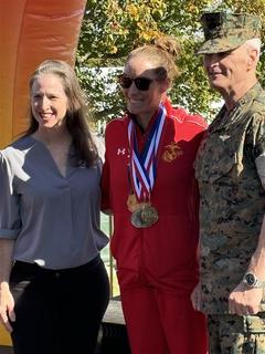 Three adults stand together smiling; the center person wears medals and a red athletic outfit, one wears a military uniform.