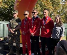 Five people pose outdoors, three in red tracksuits with medals, beside a marine and a woman, at an awards event.
