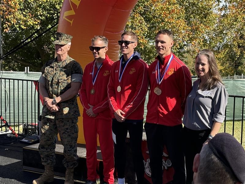 Five people pose outdoors, three in red tracksuits with medals, beside a marine and a woman, at an awards event.