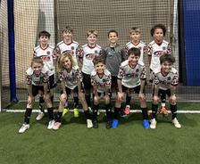 Ten boys in matching soccer uniforms pose for a team photo indoors on artificial turf, smiling at the camera.
