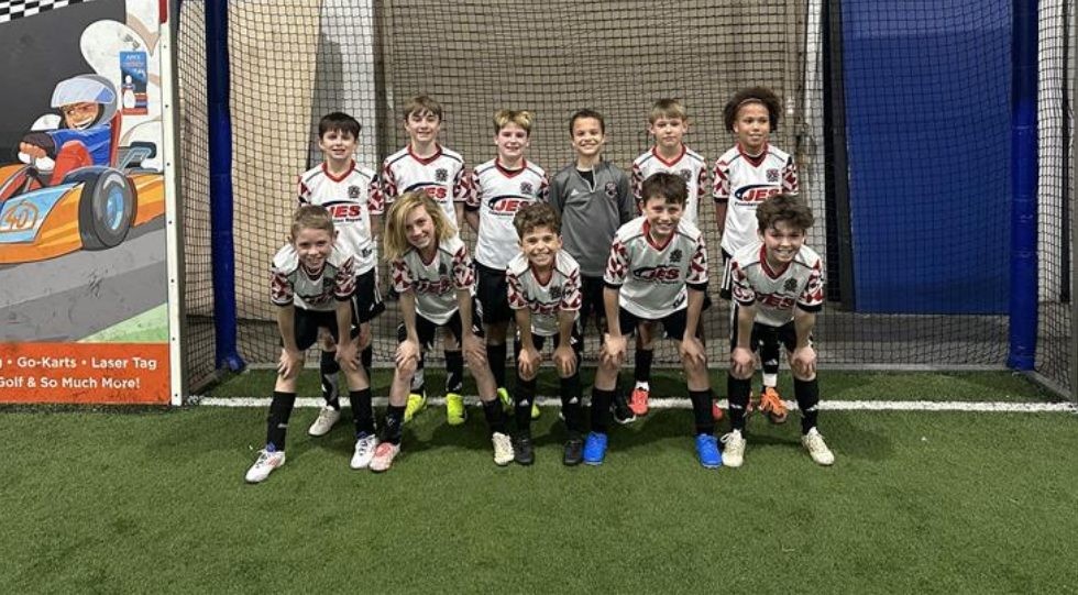 Ten boys in matching soccer uniforms pose for a team photo indoors on artificial turf, smiling at the camera.