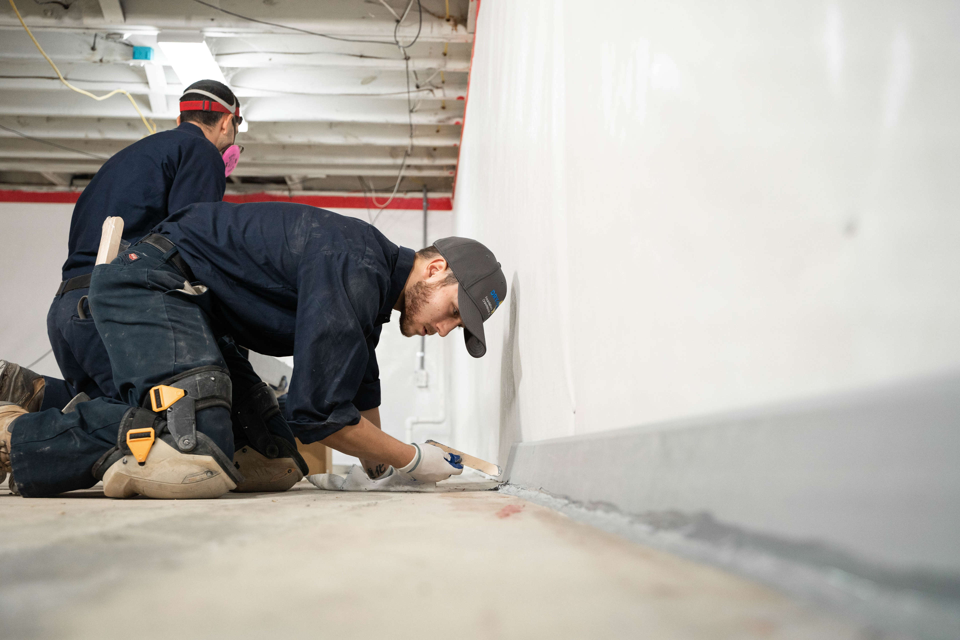 Two workers kneel on the floor, applying sealant for basement waterproofing at the base of a white wall in an unfinished room.