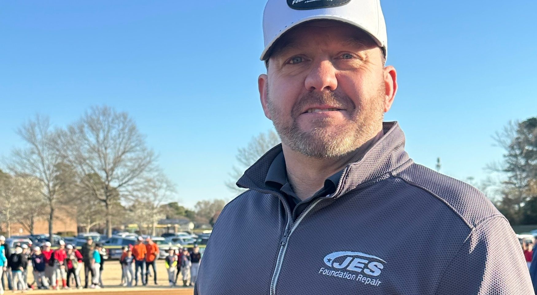 Man in JES Foundation Repair jacket and hat standing outdoors, with a crowd and trees in the background.